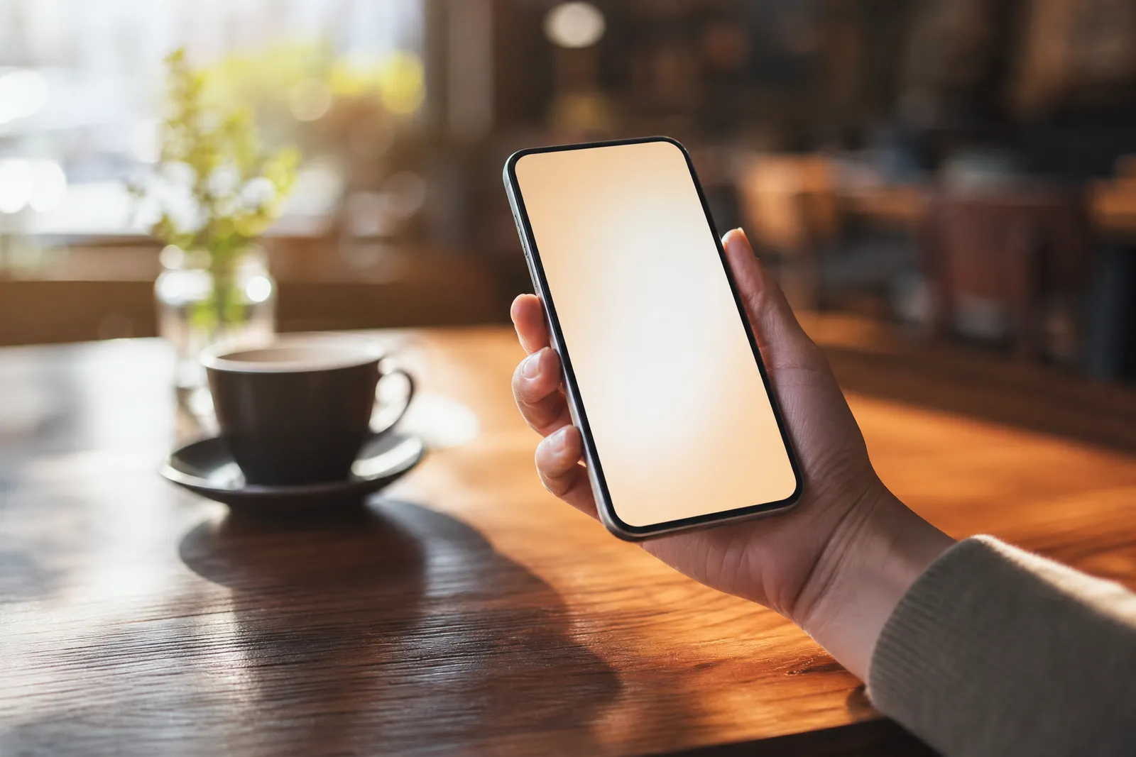 Hand holding a smartphone with a blank screen in a sunny café